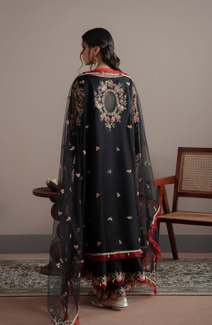 Woman wearing a black embroidered traditional outfit in a room with a chair and table.